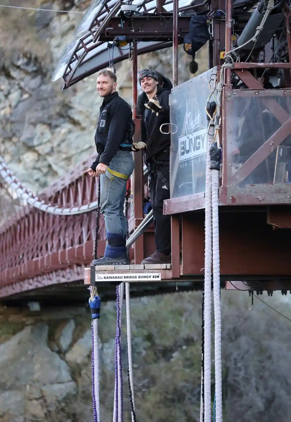 Two crew on the bridge platform beside a sign reading Kawarau Bridge Bungy 43m